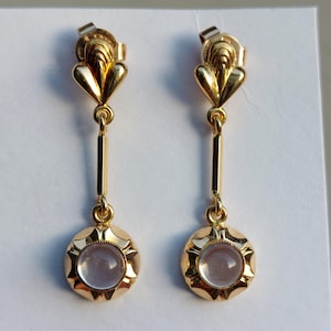 May include: A pair of gold drop earrings featuring a polished metal top section with a fan-like shape, connected by a vertical bar to a circular base. Each base holds a smooth, pale lavender cabochon stone surrounded by a scalloped metal edge.