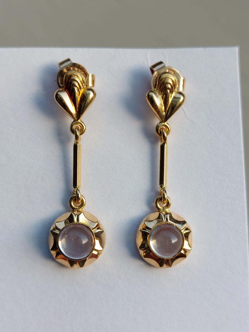 May include: A pair of gold drop earrings featuring a polished metal top section with a fan-like shape, connected by a vertical bar to a circular base. Each base holds a smooth, pale lavender cabochon stone surrounded by a scalloped metal edge.