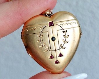 May include: A heart-shaped gold locket pendant featuring an engraved laurel wreath design, a central dark stone, and four small red stones arranged in a geometric pattern, suspended from a thin gold chain.