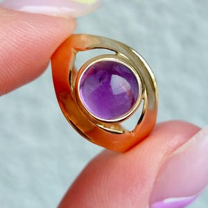 May include: A gold ring featuring a round, vibrant purple gemstone. The ring has a unique, open design with a polished finish. The gemstone is set in a bezel setting, showcasing its color and clarity. A piece of jewelry.