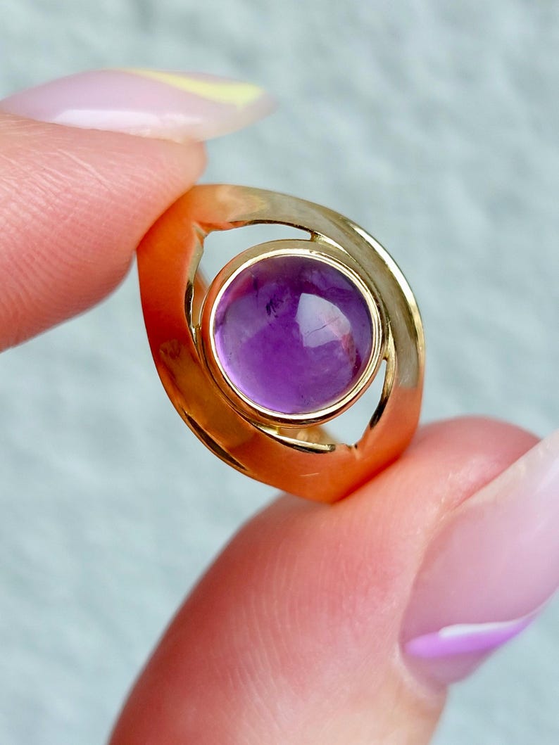 May include: A gold ring featuring a round, vibrant purple gemstone. The ring has a unique, open design with a polished finish. The gemstone is set in a bezel setting, showcasing its color and clarity. A piece of jewelry.