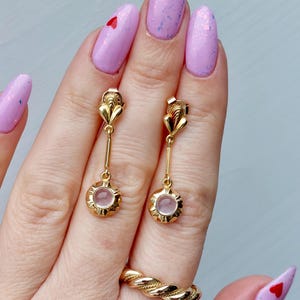 May include: A pair of gold drop earrings featuring a small, round, pale pink stone set in a scalloped gold frame, suspended from a vertical gold bar and a decorative gold top piece. A gold twisted rope-style ring is also visible on the finger.