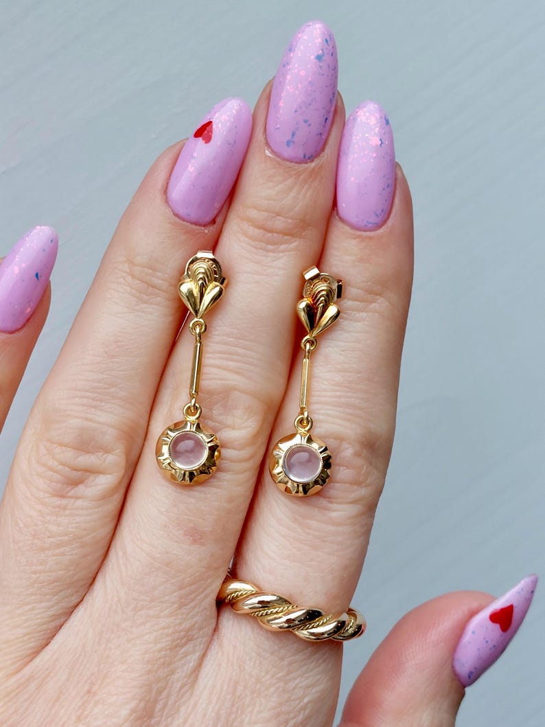 May include: A pair of gold drop earrings featuring a small, round, pale pink stone set in a scalloped gold frame, suspended from a vertical gold bar and a decorative gold top piece. A gold twisted rope-style ring is also visible on the finger.