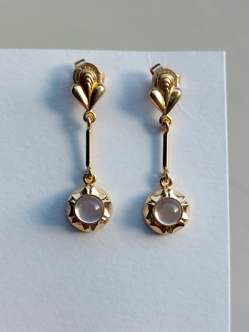 May include: A pair of gold drop earrings featuring a fan-shaped top post connected by a thin vertical bar to a circular pendant. Each pendant contains a smooth, pale pink cabochon gemstone set within a scalloped, polished gold frame.