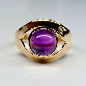 May include: A gold ring featuring a large, round, purple gemstone. The gemstone has a gradient of colors, from deep purple to lighter shades. The ring's band has a unique, open design that curves around the gemstone.