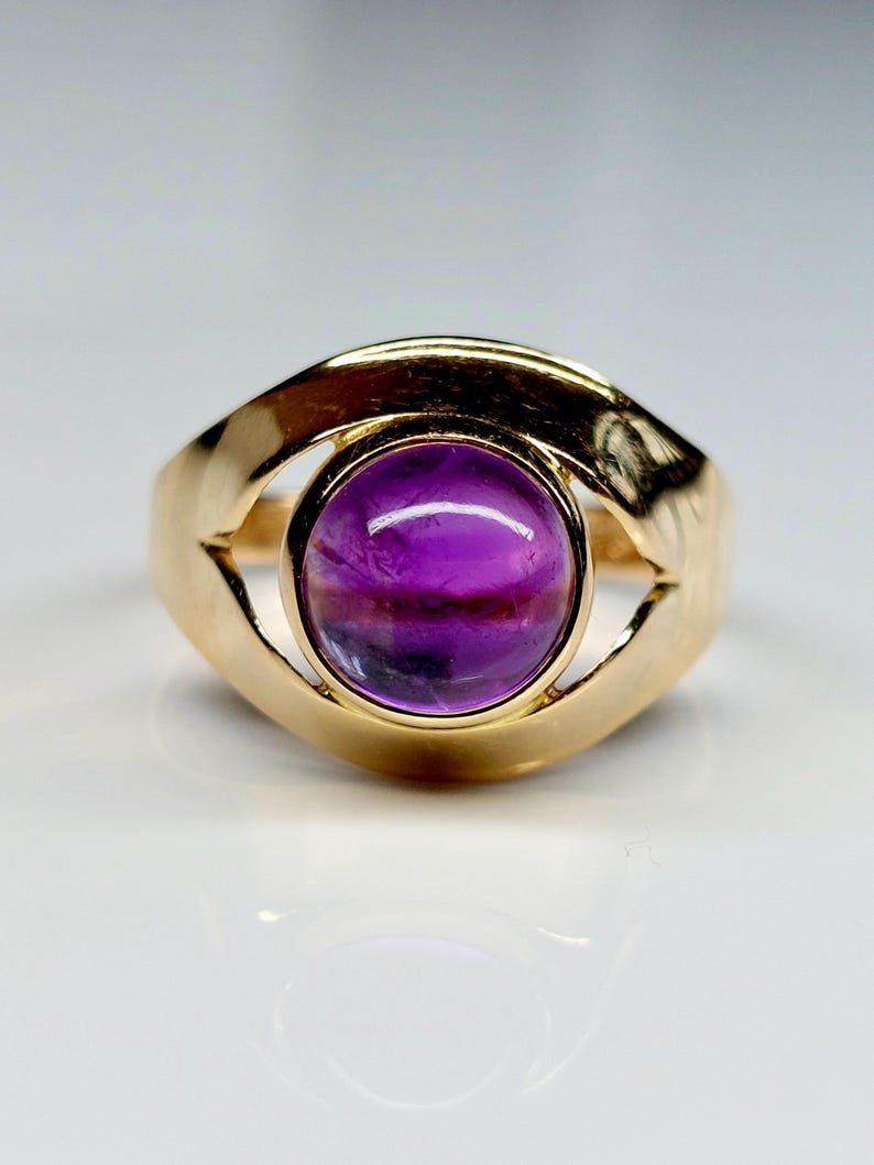May include: A gold ring featuring a large, round, purple gemstone. The gemstone has a gradient of colors, from deep purple to lighter shades. The ring's band has a unique, open design that curves around the gemstone.
