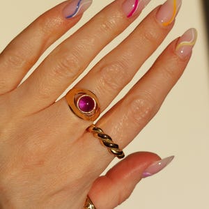 May include: A hand displaying gold rings and manicured nails. The rings include a gold signet ring with a pink gemstone, a twisted band, and a simple band. The nails are painted with a clear base and colorful wavy lines.