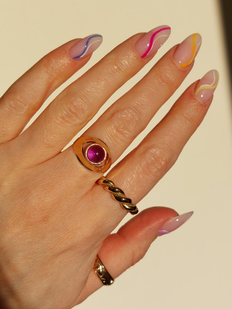 May include: A hand displaying gold rings and manicured nails. The rings include a gold signet ring with a pink gemstone, a twisted band, and a simple band. The nails are painted with a clear base and colorful wavy lines.