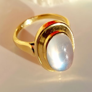 May include: A gold ring with an oval, iridescent, light blue gemstone. The ring has a simple band and a bezel setting that frames the stone. The gemstone appears to be a moonstone, and the ring is set against a soft, neutral background.