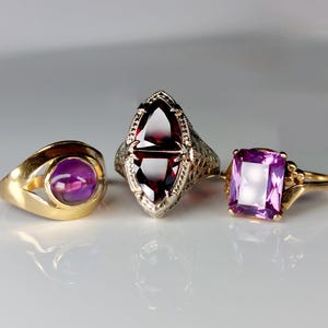 May include: Three antique rings with gemstones. A gold ring with a round purple stone, a silver ring with a maroon, elongated stone, and a gold ring with a rectangular purple stone. The rings are displayed on a white surface.