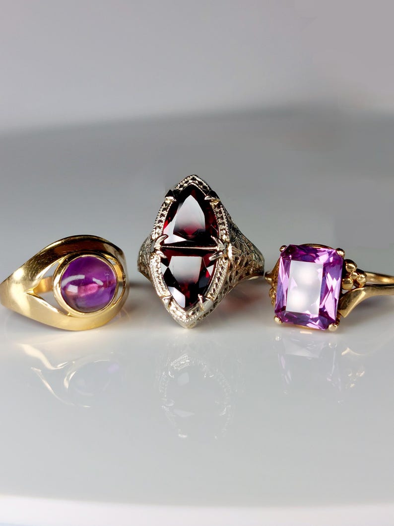 May include: Three antique rings with gemstones. A gold ring with a round purple stone, a silver ring with a maroon, elongated stone, and a gold ring with a rectangular purple stone. The rings are displayed on a white surface.