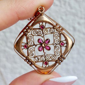 May include: A gold-toned pendant with a diamond-shaped filigree design. The pendant features a central flower and surrounding red gemstones. The intricate openwork design is accented with small, decorative details, creating a vintage aesthetic.
