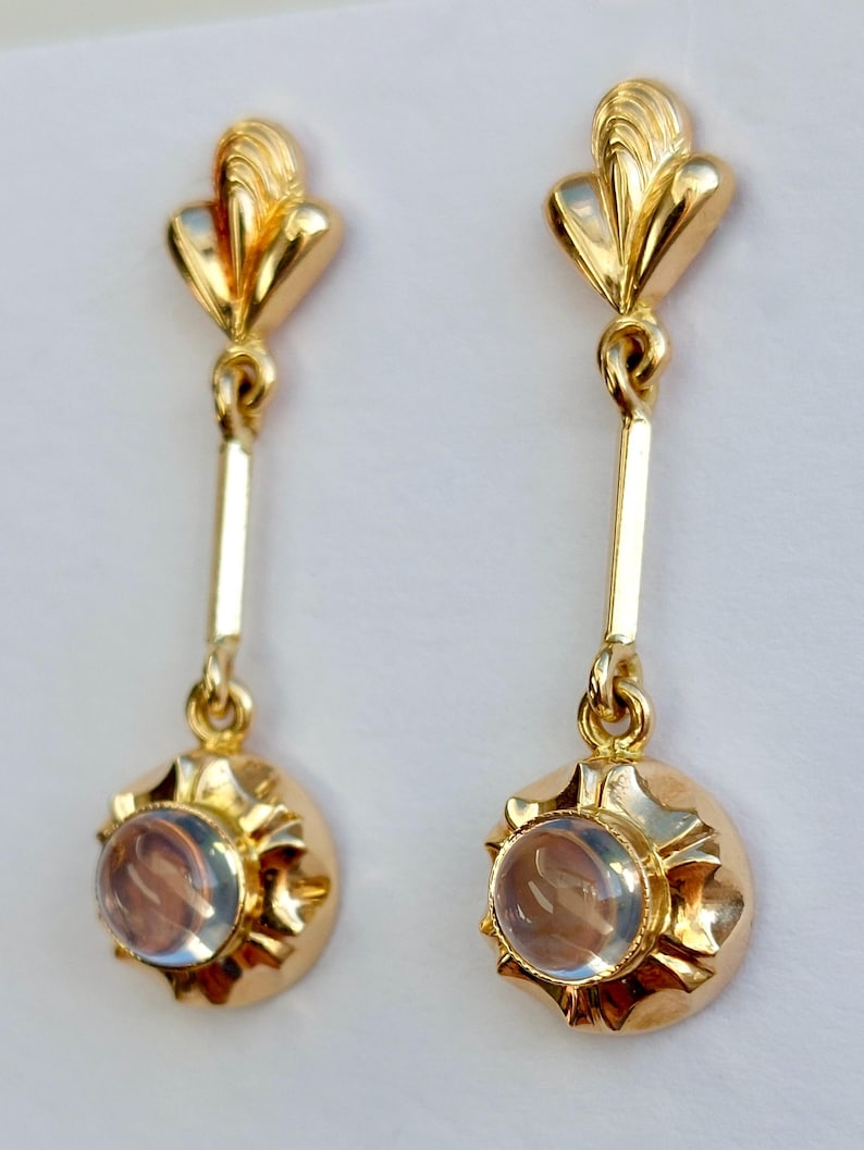 May include: A pair of gold drop earrings featuring a polished fan-shaped top post connected by a vertical bar to a circular base. Each base is set with a smooth, translucent, round cabochon stone surrounded by a scalloped, sunburst-like gold frame.