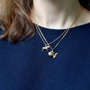 May include: Two gold necklaces with pendant charms. One necklace features a small horse pendant, and the other has a fish pendant. The necklaces are layered and worn over a dark blue top.