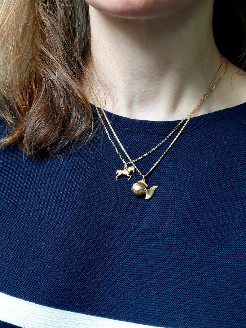 May include: Two gold necklaces with pendant charms. One necklace features a small horse pendant, and the other has a fish pendant. The necklaces are layered and worn over a dark blue top.