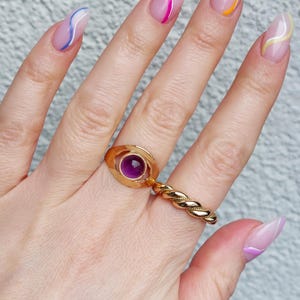 May include: A hand wearing three gold rings. One ring features a purple gemstone, another is a twisted band, and the third has an engraved design. The nails are painted with a clear base and colorful wavy lines.
