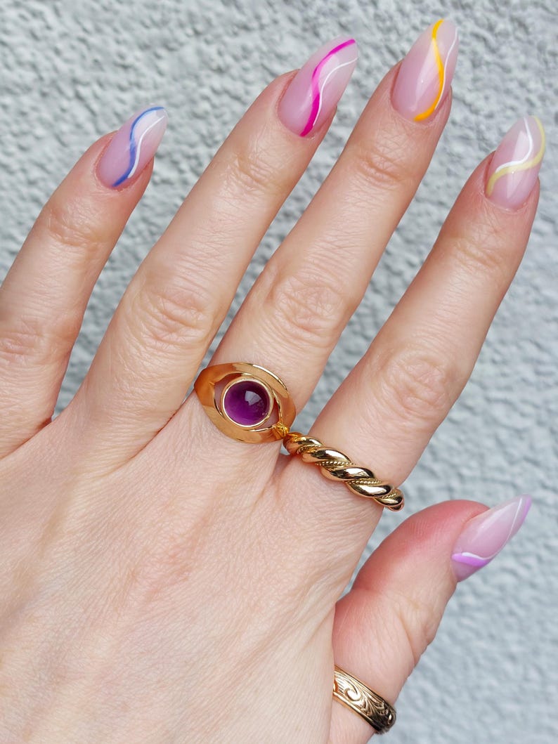 May include: A hand wearing three gold rings. One ring features a purple gemstone, another is a twisted band, and the third has an engraved design. The nails are painted with a clear base and colorful wavy lines.
