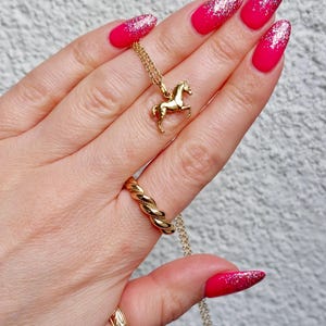 May include: A gold-colored horse pendant necklace with a matching twisted ring and a patterned ring. The nails are painted with a bright pink color and glitter.
