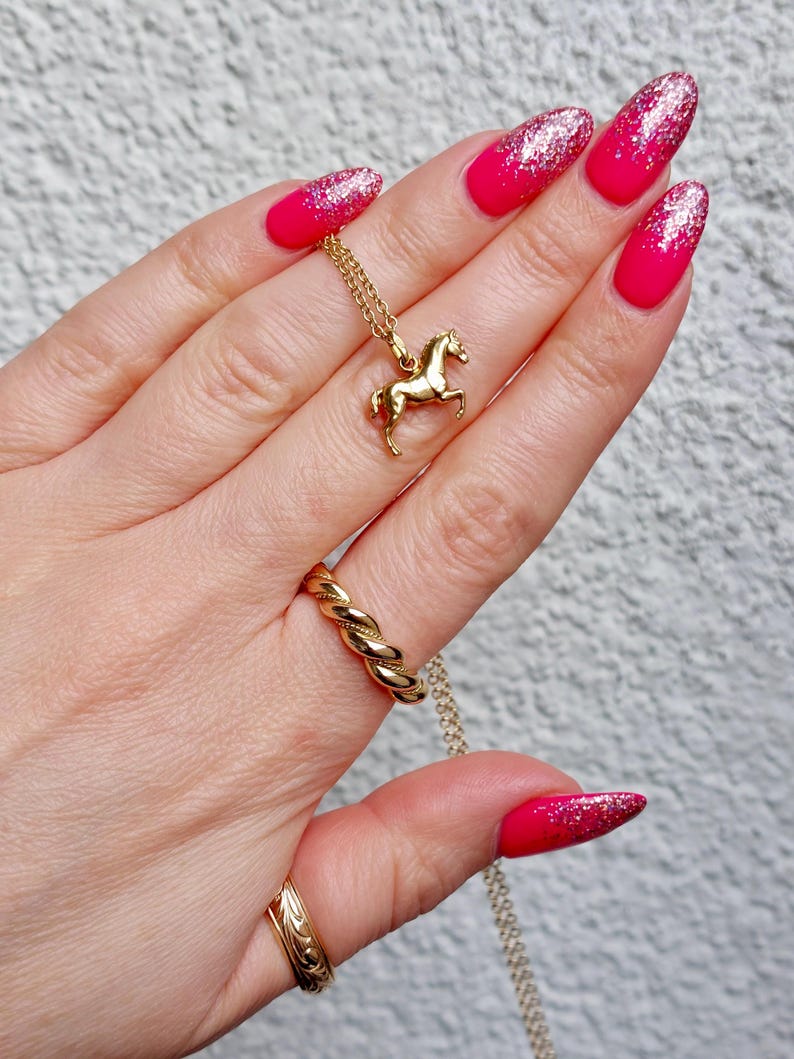 May include: A gold-colored horse pendant necklace with a matching twisted ring and a patterned ring. The nails are painted with a bright pink color and glitter.