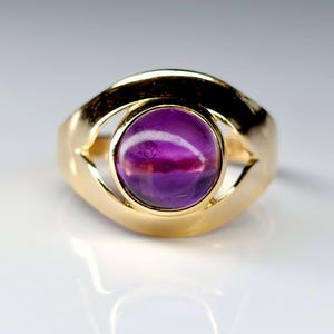 May include: A gold ring featuring a large, oval-shaped, purple gemstone. The ring's band is a thick, polished gold, with an open design around the gemstone. The gemstone has a polished, smooth surface, reflecting light.