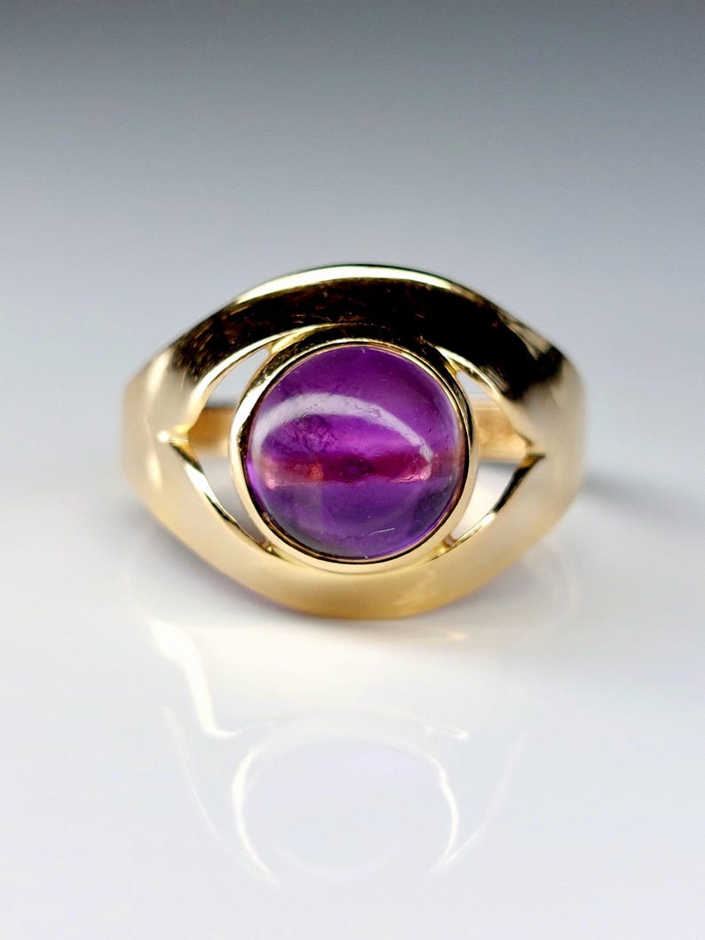 May include: A gold ring featuring a large, oval-shaped, purple gemstone. The ring's band is a thick, polished gold, with an open design around the gemstone. The gemstone has a polished, smooth surface, reflecting light.