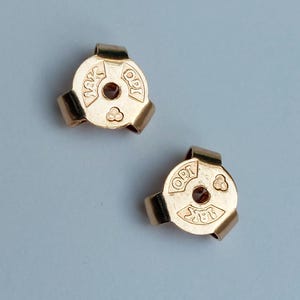 May include: Two gold-colored earring backs with a circular design and three curved prongs. Each piece features the stamped text 18K and OPI, along with a small triangular symbol consisting of three circles, surrounding a central hole for a post.