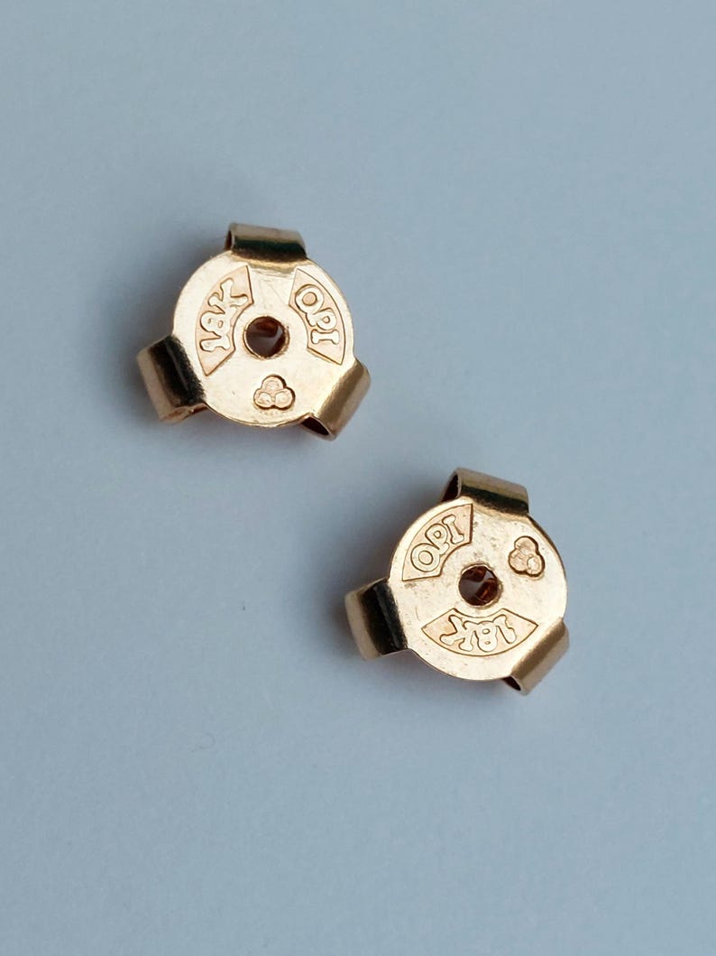 May include: Two gold-colored earring backs with a circular design and three curved prongs. Each piece features the stamped text 18K and OPI, along with a small triangular symbol consisting of three circles, surrounding a central hole for a post.