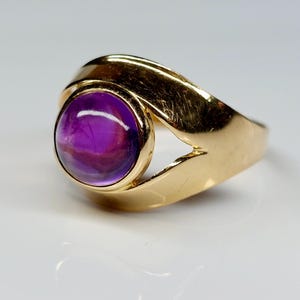 May include: A gold ring featuring a large, round, purple gemstone. The ring's band has a unique, flowing design that curves around the gemstone. The gemstone is a deep purple color with some lighter purple and white marbling.
