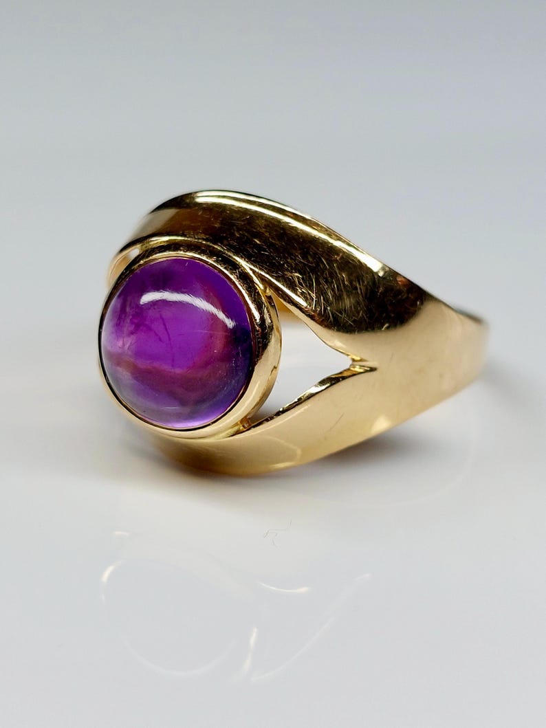 May include: A gold ring featuring a large, round, purple gemstone. The ring's band has a unique, flowing design that curves around the gemstone. The gemstone is a deep purple color with some lighter purple and white marbling.