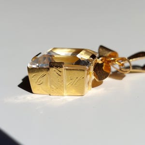Op de afbeelding: Een goudkleurige hanger met een heldere, rechthoekige edelsteen. De hanger heeft een strikvormige balein en gegraveerde letters aan de zijkanten. De edelsteen is facetgeslepen en reflecteert licht. Het ontwerp is elegant en luxueus.