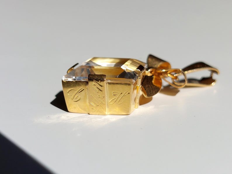 Op de afbeelding: Een goudkleurige hanger met een heldere, rechthoekige edelsteen. De hanger heeft een strikvormige balein en gegraveerde letters aan de zijkanten. De edelsteen is facetgeslepen en reflecteert licht. Het ontwerp is elegant en luxueus.