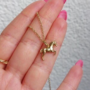 May include: A gold-colored horse pendant necklace. The horse is in a trotting pose, and the necklace chain is delicate. The pendant is held in a hand with pink nail polish.