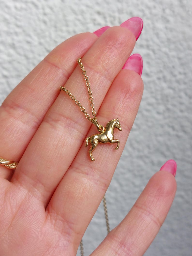 May include: A gold-colored horse pendant necklace. The horse is in a trotting pose, and the necklace chain is delicate. The pendant is held in a hand with pink nail polish.