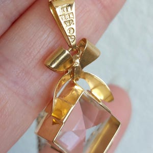 Op de afbeelding: Gouden hangerketting met een heldere, facetgeslepen, vierkante kristal. De hanger heeft een gouden strik ontwerp en hangt aan een fijne ketting. De hanger heeft een gouden balein met markeringen.