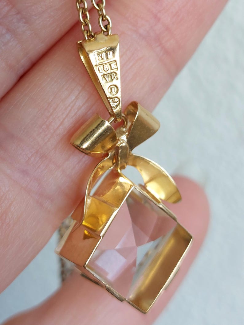 Op de afbeelding: Gouden hangerketting met een heldere, facetgeslepen, vierkante kristal. De hanger heeft een gouden strik ontwerp en hangt aan een fijne ketting. De hanger heeft een gouden balein met markeringen.