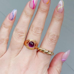 May include: A hand displaying a gold ring with a purple gemstone and a twisted gold band ring. The nails are long and painted with a clear base and colorful wavy lines in pink, yellow, and blue.