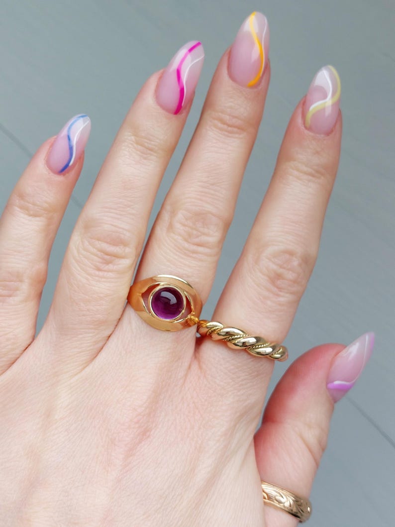 May include: A hand displaying a gold ring with a purple gemstone and a twisted gold band ring. The nails are long and painted with a clear base and colorful wavy lines in pink, yellow, and blue.