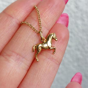 May include: A gold-colored horse pendant necklace. The pendant is a detailed miniature of a horse in a trotting pose. The necklace chain is delicate and also gold-colored. The background is a soft, neutral color.