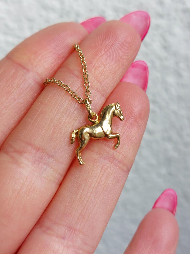 May include: A gold-colored horse pendant necklace. The pendant is a detailed miniature of a horse in a trotting pose. The necklace chain is delicate and also gold-colored. The background is a soft, neutral color.