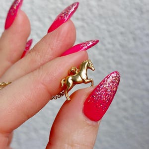 May include: A gold-colored horse pendant necklace. The horse is in a rearing pose. The background is a hand with long, bright pink nails with glitter accents.