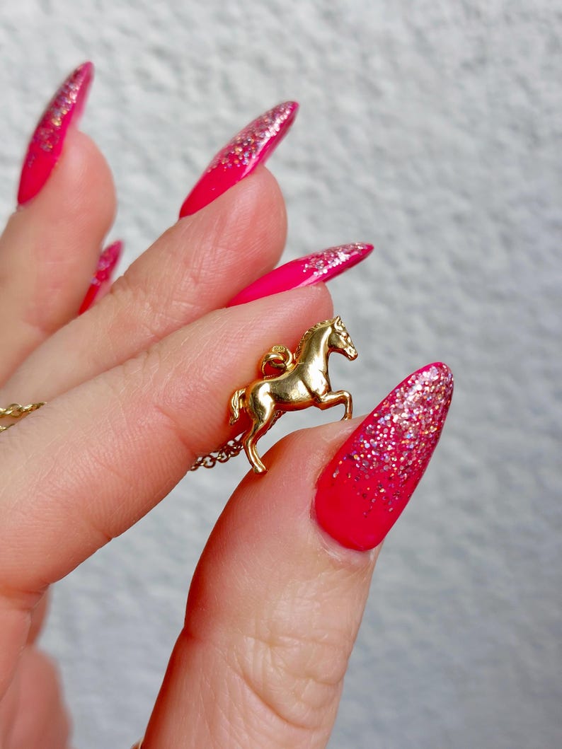 May include: A gold-colored horse pendant necklace. The horse is in a rearing pose. The background is a hand with long, bright pink nails with glitter accents.