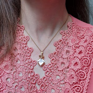 Op de afbeelding: Een goudkleurige ketting met een vierkante, heldere hanger en een klein strikje. De ketting wordt gedragen over een roze top met een gedetailleerde kanten halslijn. De afbeelding is een close-up van de ketting en de top.