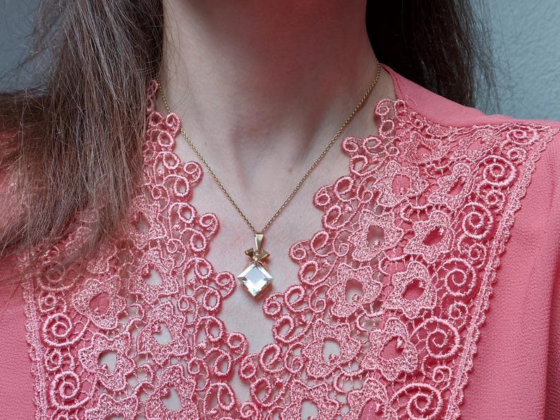 Op de afbeelding: Een goudkleurige ketting met een vierkante, heldere hanger en een klein strikje. De ketting wordt gedragen over een roze top met een gedetailleerde kanten halslijn. De afbeelding is een close-up van de ketting en de top.