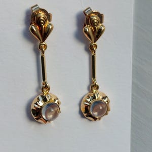 May include: A pair of gold drop earrings featuring a fan-shaped top post connected to a vertical bar and a circular base. Each earring is set with a small, translucent, round cabochon stone that displays a soft, iridescent blue glow.