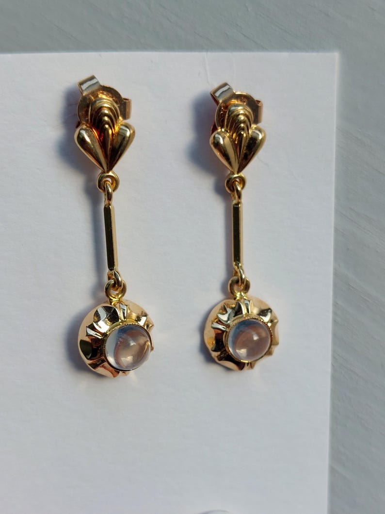 May include: A pair of gold drop earrings featuring a fan-shaped top post connected to a vertical bar and a circular base. Each earring is set with a small, translucent, round cabochon stone that displays a soft, iridescent blue glow.