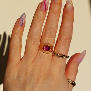 May include: Hand with long, almond-shaped nails, each painted with a clear base and a colorful, wavy line design. A gold ring with a large, oval, pink gemstone and two other gold rings adorn the fingers.
