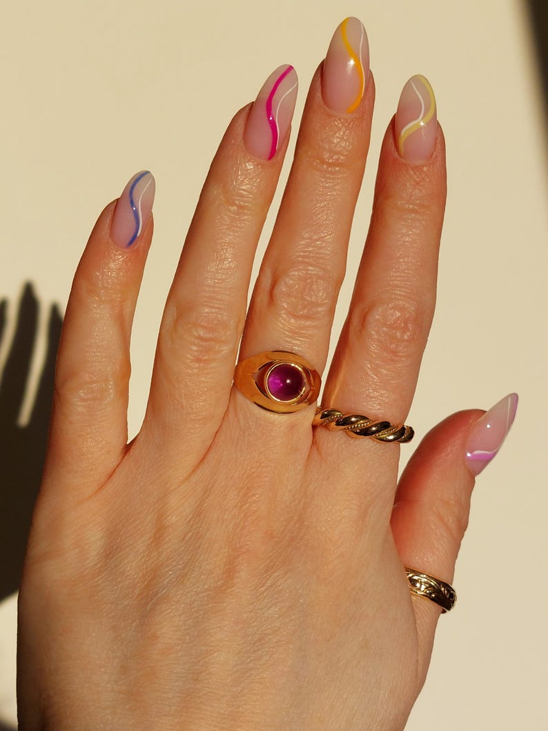 May include: Hand with long, almond-shaped nails, each painted with a clear base and a colorful, wavy line design. A gold ring with a large, oval, pink gemstone and two other gold rings adorn the fingers.