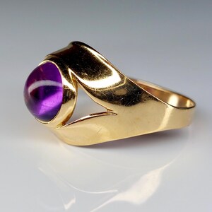 May include: A gold ring featuring a large, round, deep purple gemstone. The ring's band has a unique, open design with a polished finish. The gemstone is set in a bezel setting, showcasing its color and shape.