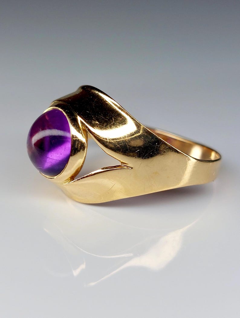 May include: A gold ring featuring a large, round, deep purple gemstone. The ring's band has a unique, open design with a polished finish. The gemstone is set in a bezel setting, showcasing its color and shape.