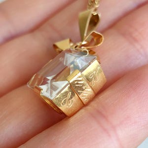 Op de afbeelding: Een goudkleurige hangerketting met een heldere, rechthoekige kristal. De kristal is gezet in een gouden frame met een strikvormige balein. Het gouden frame heeft gegraveerde details. De ketting wordt in een hand gehouden.